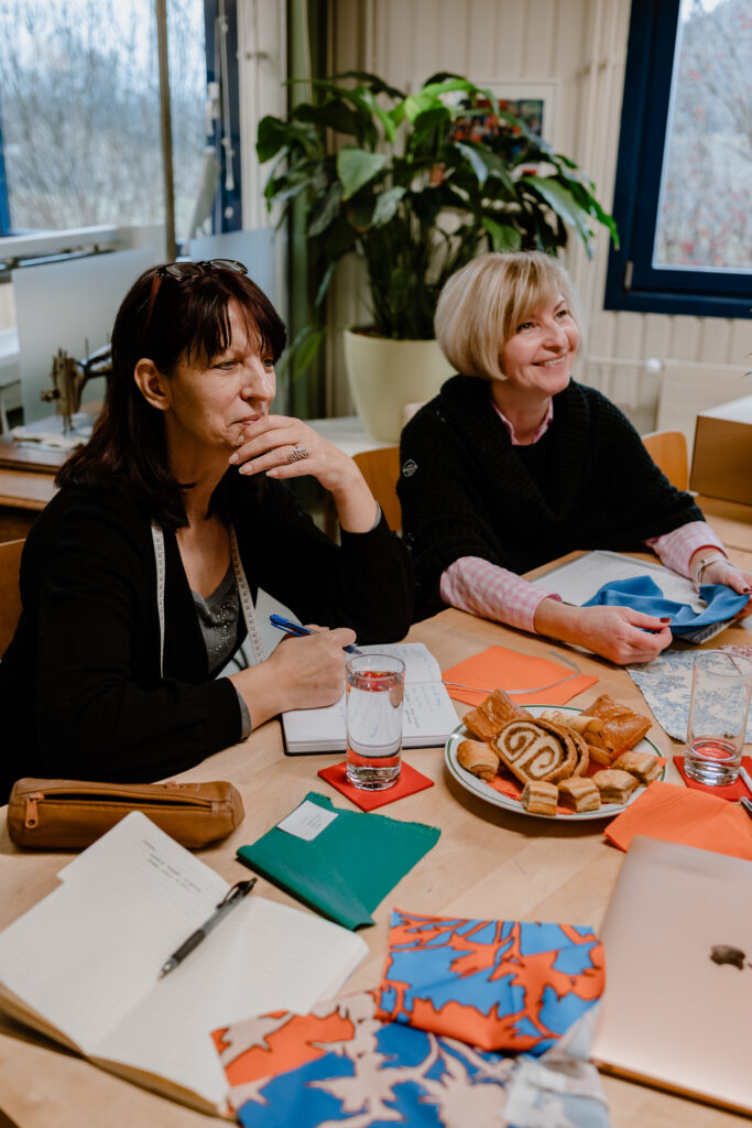 Two women sit together at a wooden meeting table in conversation, with colourful fabric swatches in a bold red, orange and blue botanical print spread across the table alongside notebooks, pastries, and glasses of water. A large plant and winter trees are visible through the window behind them, suggesting a relaxed but productive design or production meeting.