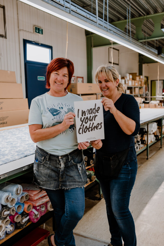 Two female garment factory workers standing together holding a sign reading "I made your clothes" in front of fabric rolls and cutting tables