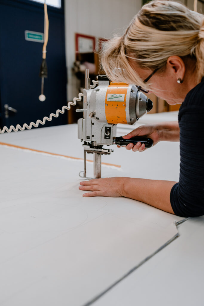 A blonde woman wearing glasses leans over a large white cutting table, operating a handheld industrial rotary cutting machine to cut fabric. Shot in a professional garment production environment with equipment visible in the background.