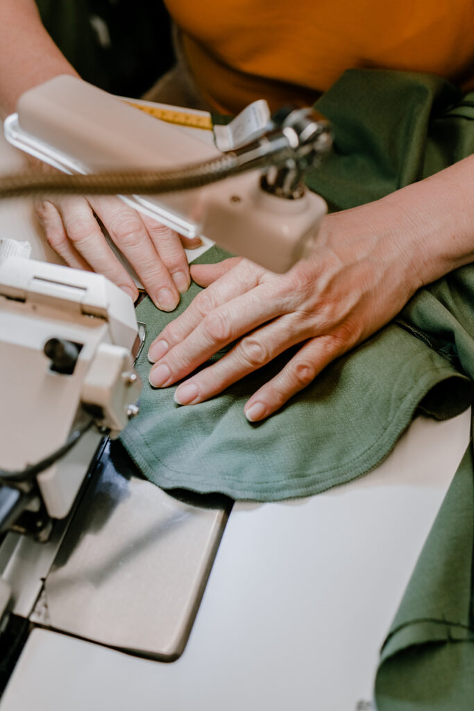 A tight close-up shot of a person's hands carefully feeding green fabric through the needle of an industrial sewing machine, highlighting skilled garment construction and hands-on craftsmanship.