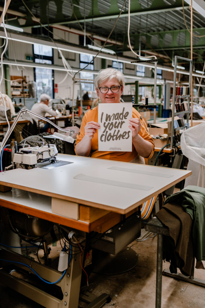 Female garment worker seated at an industrial sewing machine on the factory floor, holding a sign reading "I made your clothes"