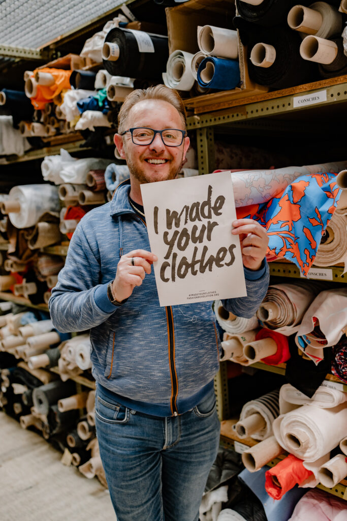 Male garment factory worker standing in front of shelving stacked with colourful fabric rolls, holding a sign reading "I made your clothes".