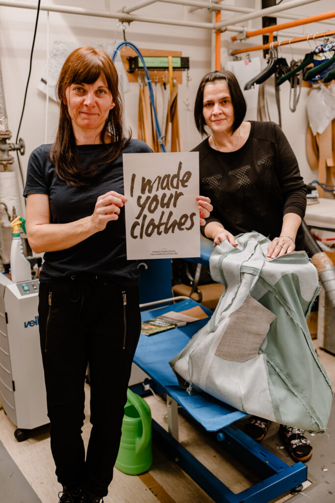 Two female garment workers standing at a pressing station holding a sign reading "I made your clothes".