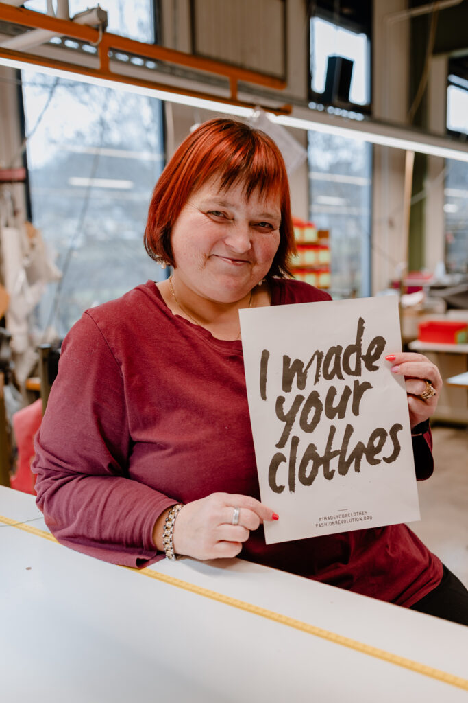 Female garment worker in a red top seated at a cutting table, holding a sign reading "I made your clothes".