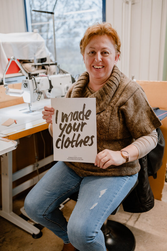 Female garment worker seated at a workstation, smiling and holding a sign reading "I made your clothes"