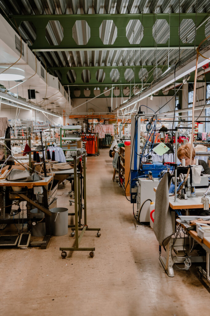 A long industrial garment factory floor lined with rows of sewing machines and workstations stretching into the distance, with green decorative ceiling panels, hanging garments, and equipment visible throughout the well-lit production space.