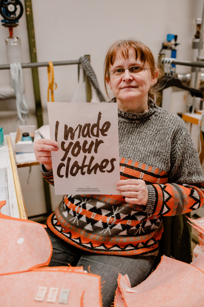 Female garment worker in a patterned jumper seated among fabric, holding a sign reading "I made your clothes".