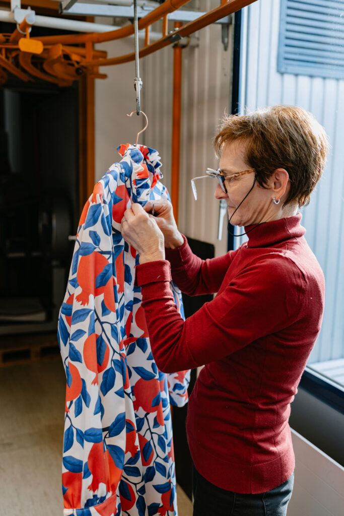 A woman wearing glasses and a red turtleneck sweater carefully inspects a garment hanging from an overhead rail, featuring a striking large-scale botanical print in red, white and blue with fruit and leaf motifs. Shot in a bright studio or factory space with natural window light.