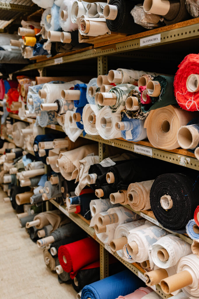 Tall wooden shelving units packed tightly with bolts of rolled fabric in a wide range of colours including red, blue, navy, cream, beige, and black. Fabric rolls are stored both horizontally and upright, suggesting a large-scale manufacturing or textile storage facility.
