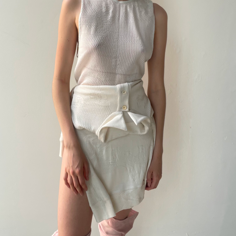 Model wearing a cream sleeveless knit top with subtle texture, paired with an ivory asymmetric wrap mini skirt featuring button fastenings and a folded ruffle panel at the hip. The skirt has a jacquard or embossed fabric finish. Partially visible pink shoes at the hem.