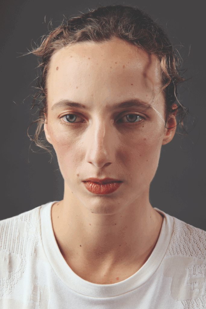 Close-up portrait shot of a model with tousled brown hair and freckled skin, wearing a white seamless textured crew-neck top. Bold rust-red lip colour. Neutral, direct expression against a dark grey background.