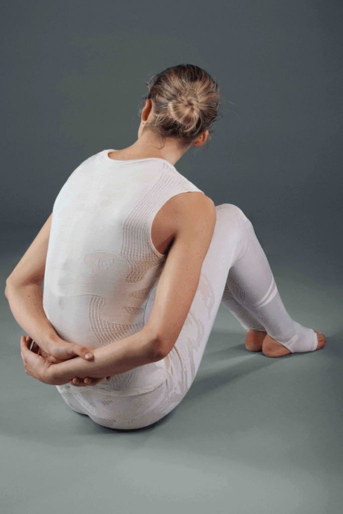 Rear view of a model seated on a grey studio floor, wearing a white seamless knit sleeveless bodysuit with subtle tonal pattern detailing, paired with white seamless leggings. Hair in a loose bun.