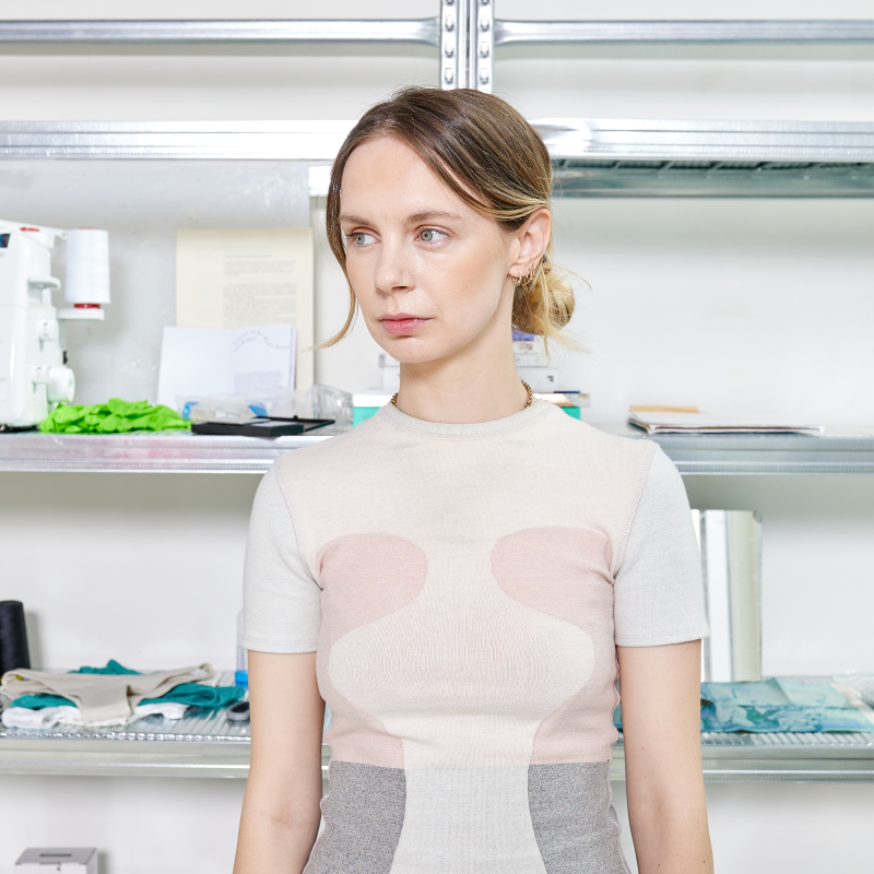 Model wearing a fitted short-sleeve knit top in cream and grey colour-blocking, with soft pink oval panel detailing across the chest. Hair styled in a loose bun with gold hoop earrings. Shot in front of metal shelving in a fashion studio or atelier environment.