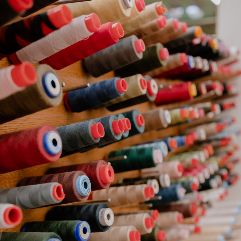 Closely packed rows of industrial thread spools in a wide range of colours — including reds, navy, teal, forest green, beige, taupe, and grey — stored horizontally on a wooden wall-mounted rack. Many spools have red plastic end caps. Shot at an angle with shallow depth of field, evoking a professional manufacturing or textile production environment.