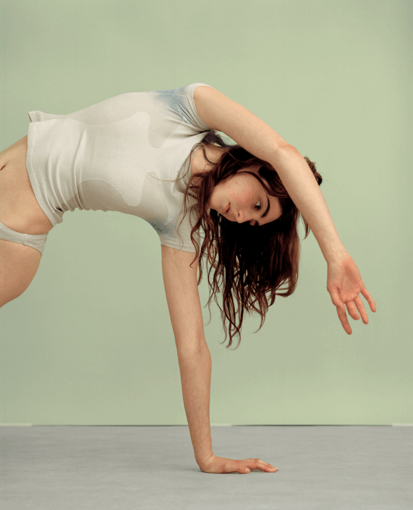 Model performing a deep lateral backbend, one hand touching the floor and the other arm arching overhead, long dark hair falling loose. Wearing a white seamless sleeveless vest top with soft cloud-like tonal print and matching white briefs. Shot against a muted sage green wall with grey floor, emphasising movement and body in motion.