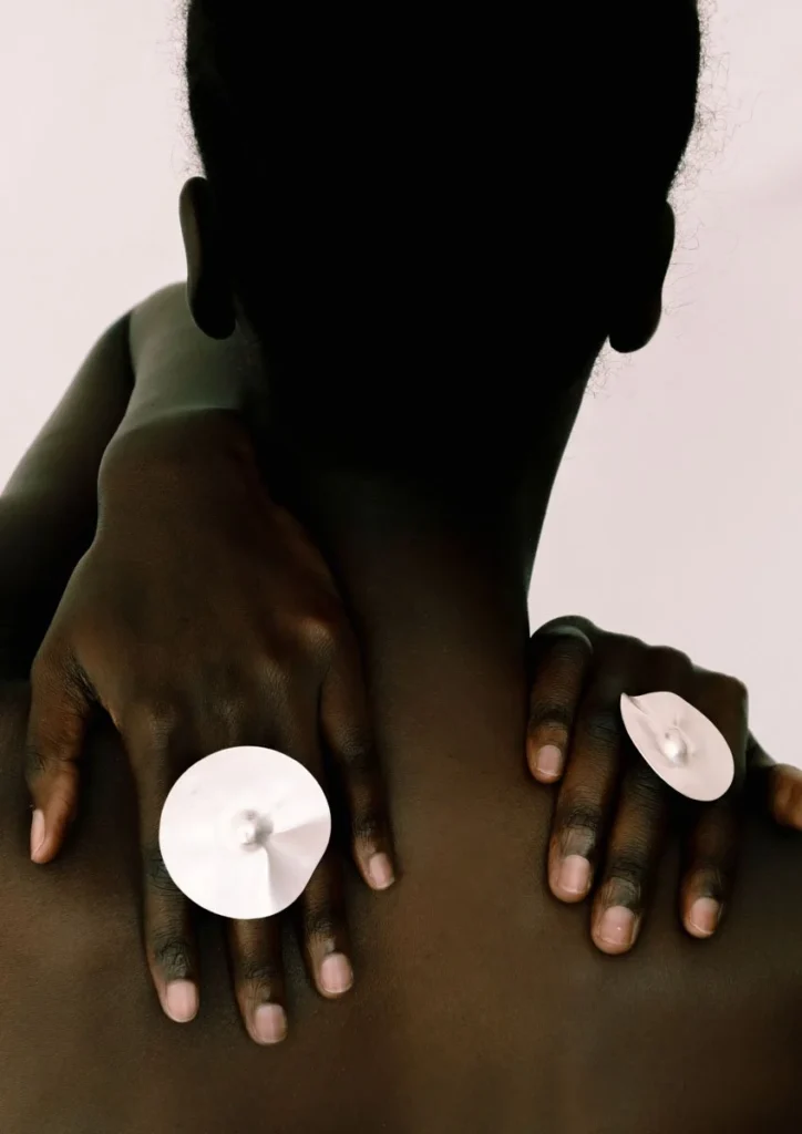 Both hands of a model rest against a bare back, each wearing an oversized circular flat disc ring in silver with a small central raised detail. The upper body and head are rendered as a dark silhouette against a soft pale grey-white background, creating a dramatic high-contrast editorial composition that draws focus to the sculptural jewellery.