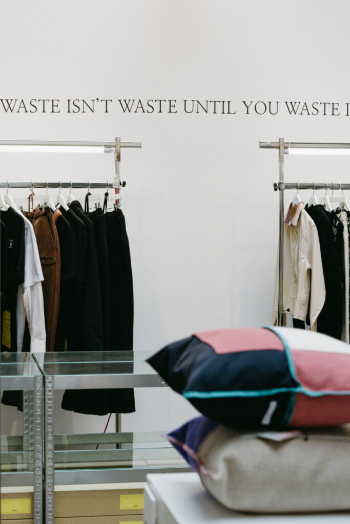 A minimalist retail space displaying clothing racks with dark and neutral garments, alongside stacked textile cushions in a patchwork design, with the phrase 'Waste Isn't Waste Until You Waste It' written on the wall.
